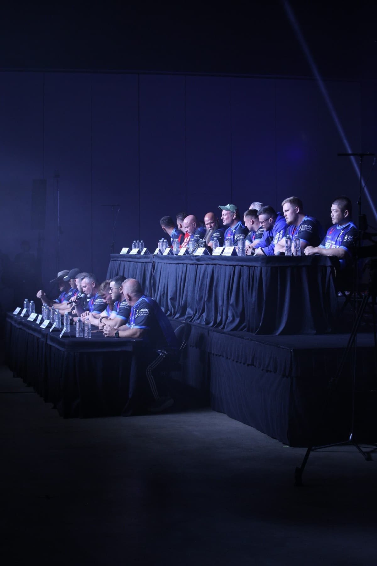 East vs West 24 press day: the full armwrestling athlete panel seated at two tiers of tables under a single shaft of light
