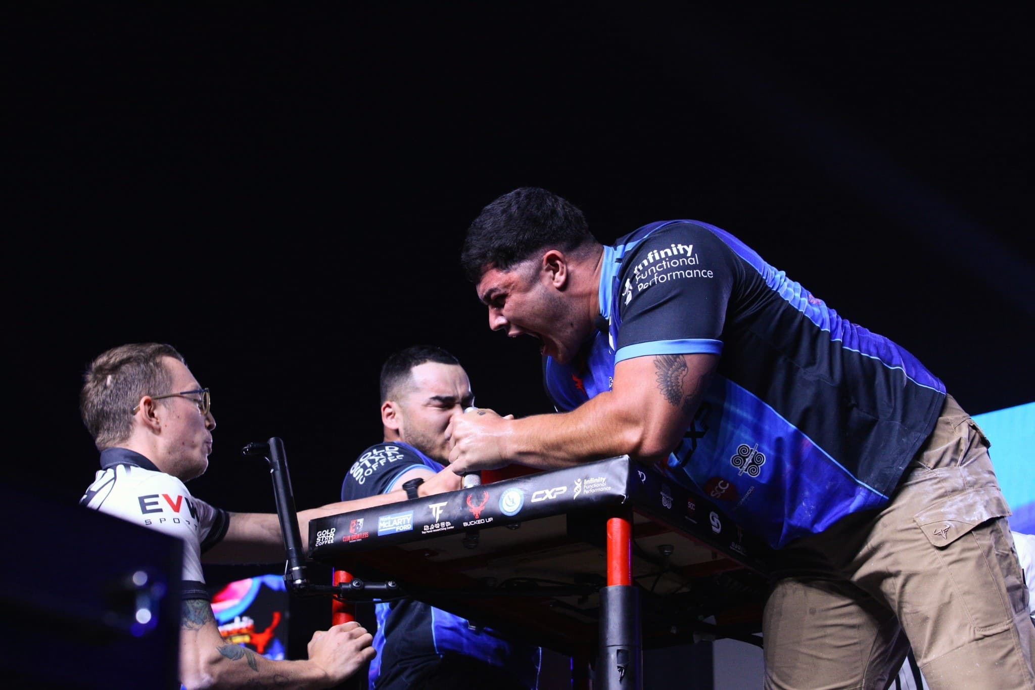 Armwrestling match at East vs West 24: a competitor roars mid-pull while the referee watches the elbow pad