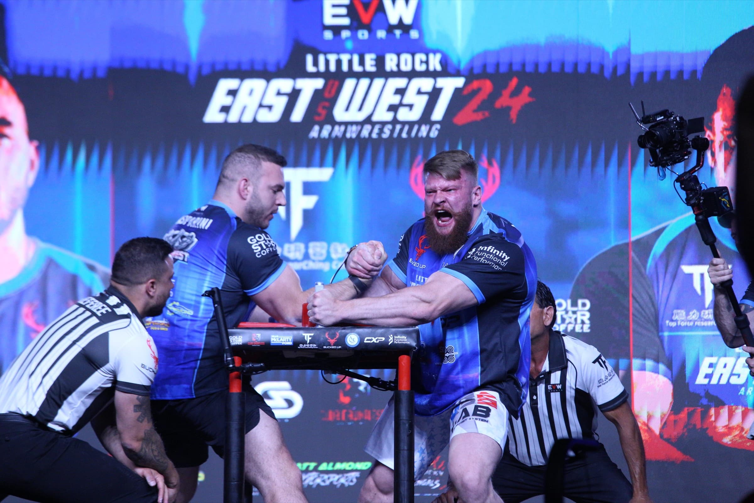 Armwrestling at East vs West 24: two competitors locked up at the table as the referees crouch to watch the pin pad