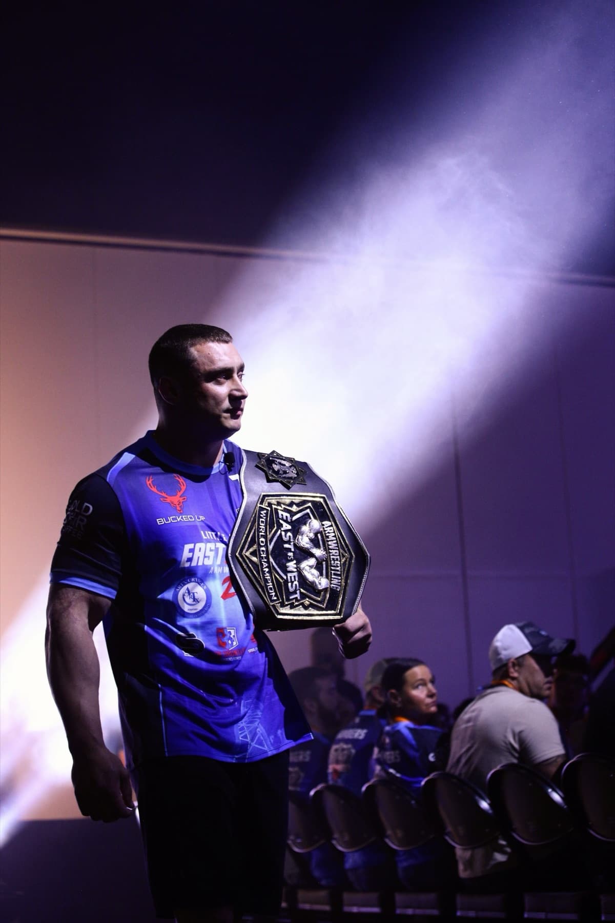 East vs West 24: an armwrestling world champion walks out through the haze holding the title belt
